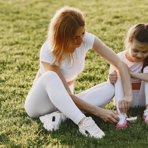 A mother and daughter in activewear bond outdoors on a sunny day, tying shoelaces on the grass.