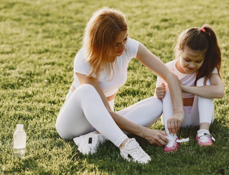A mother and daughter in activewear bond outdoors on a sunny day, tying shoelaces on the grass.